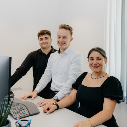 ausbildung-schicker-steuerkanzlei Drei Auszubildende stehen gemeinsam an einem Schreibtisch und lächeln in die Kamera.