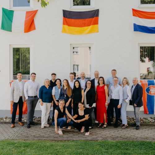 steuerkanzlei-schicker-augsburg Das Team der Steuerkanzlei Schicker und Kollegen steht vor dem Bürogebäude und an den Fenstern sind Flaggen von Italien, Deutschland und Frankreich angebracht.